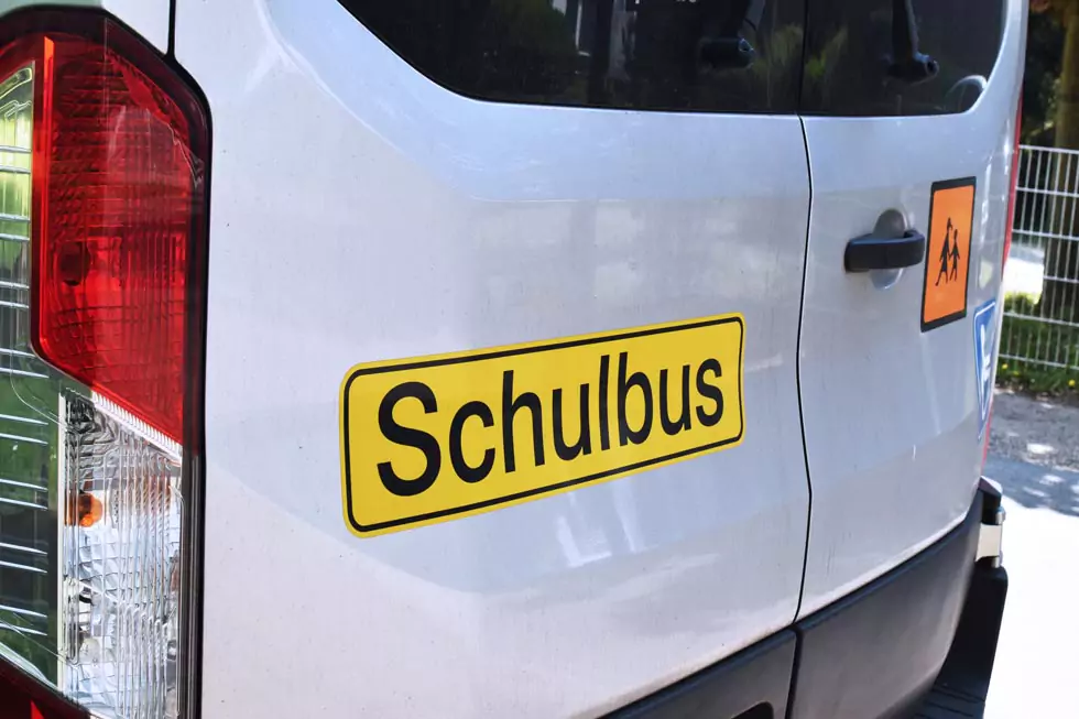 Hinteransicht eines weißen Kleinbus mit gelben Aufkleber Schulbus und Orange/schwarzen Icon mit 2 Kindern