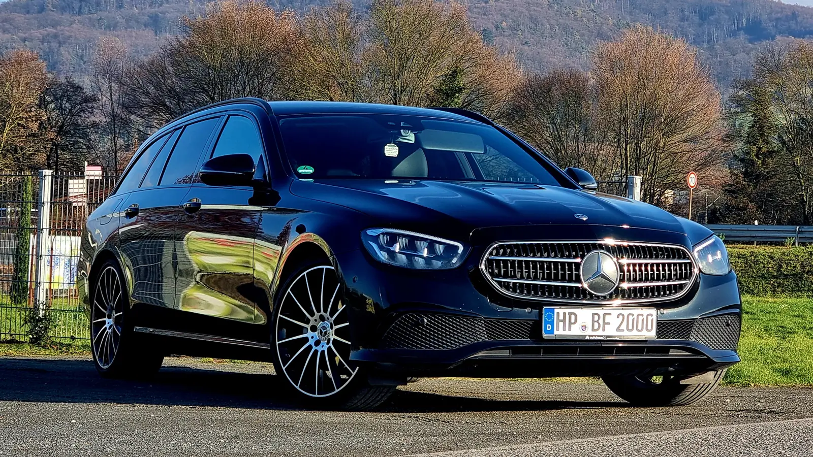 Schwarzer Mercedes E-Klasse Kombi mit markantem Kühlergrill und großen Felgen vor bewaldetem Hügel im Hintergrund